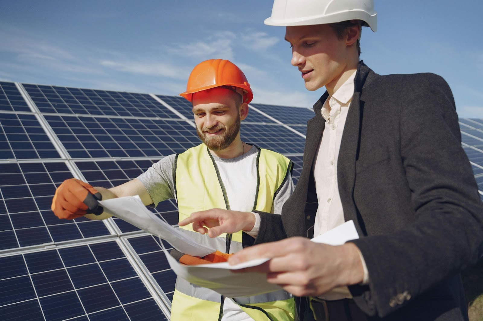 Two men next to a solar array wearing hard hats and looking at paperwork